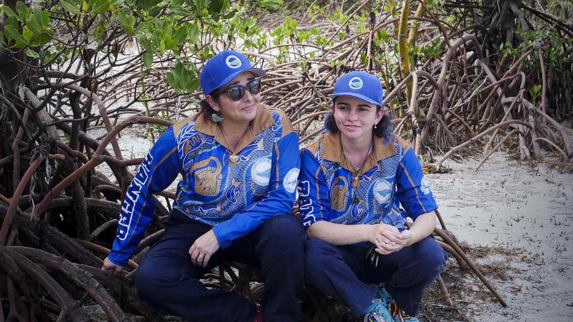 Two women in blue rangers clothing sit on a piece of wood b some mangroves.