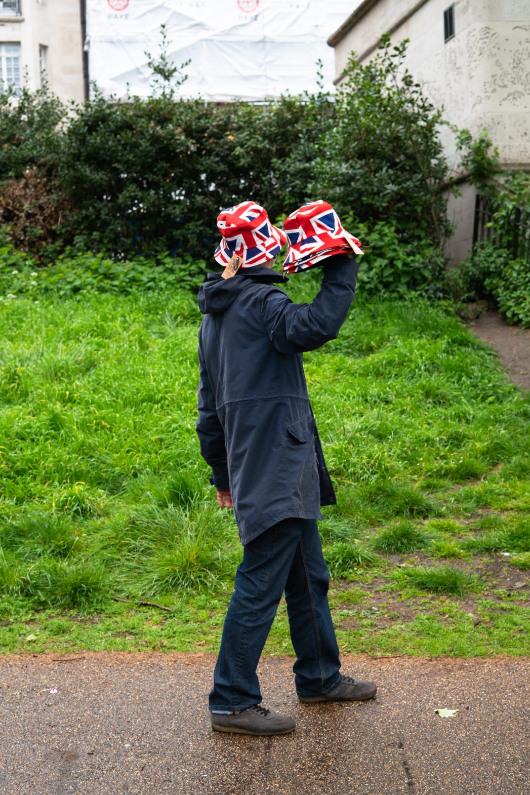 A man in a dark jacket and trousers walking, wearing a Union Jack hat and holding up others to sell.
