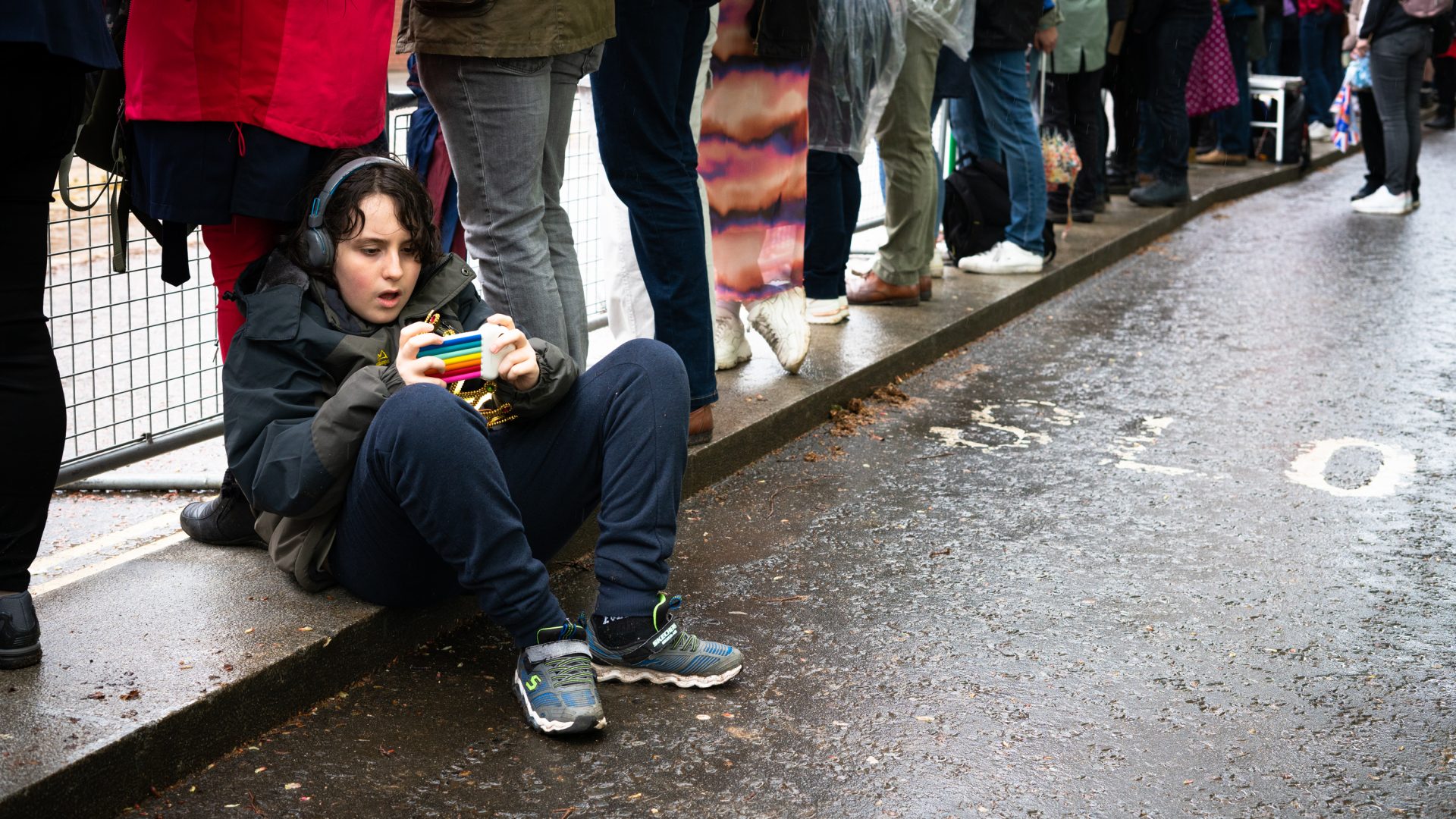 A child sits on the kerb with his phone against a backdrop of queuers.