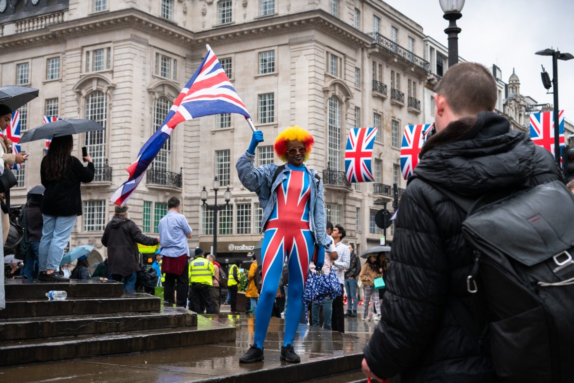 A reveller wearing a Union Jack skinsuit and a red-and-yellow wig holding a flag in Piccadilly Circus.