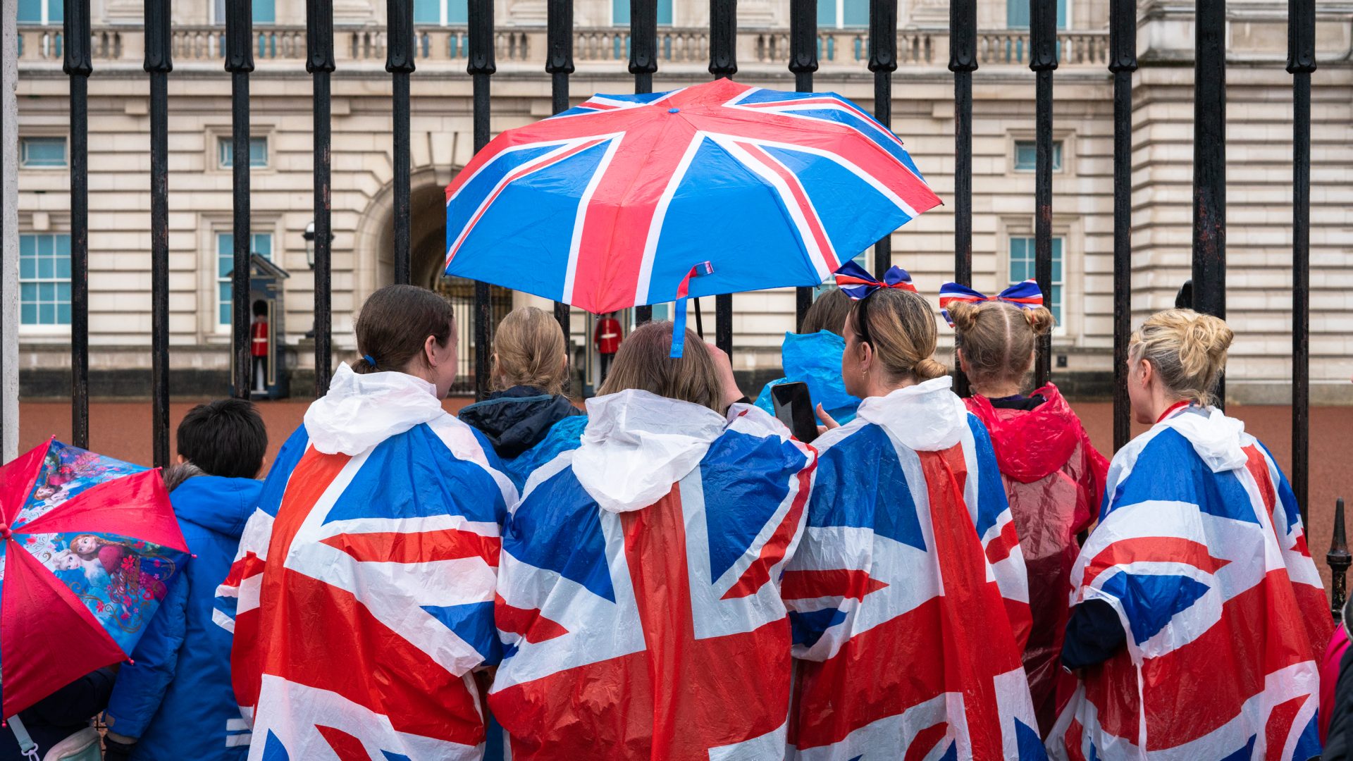A small crowd wearing Union Jack capes and holding a Union Jack umbrella in front of the gates at Buckingham Palace.