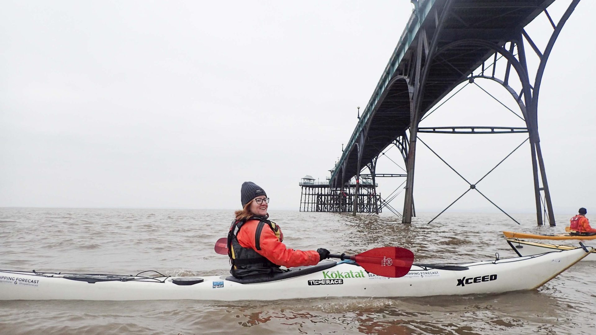 A woman looks to the camera and smiles as she pauses her kayak below a bridge.