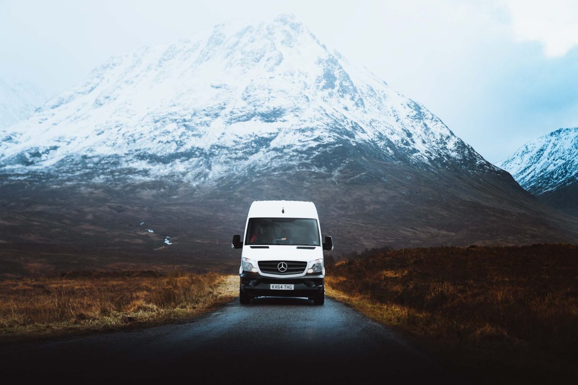 A white camper van drives away from some snow covered mountains.