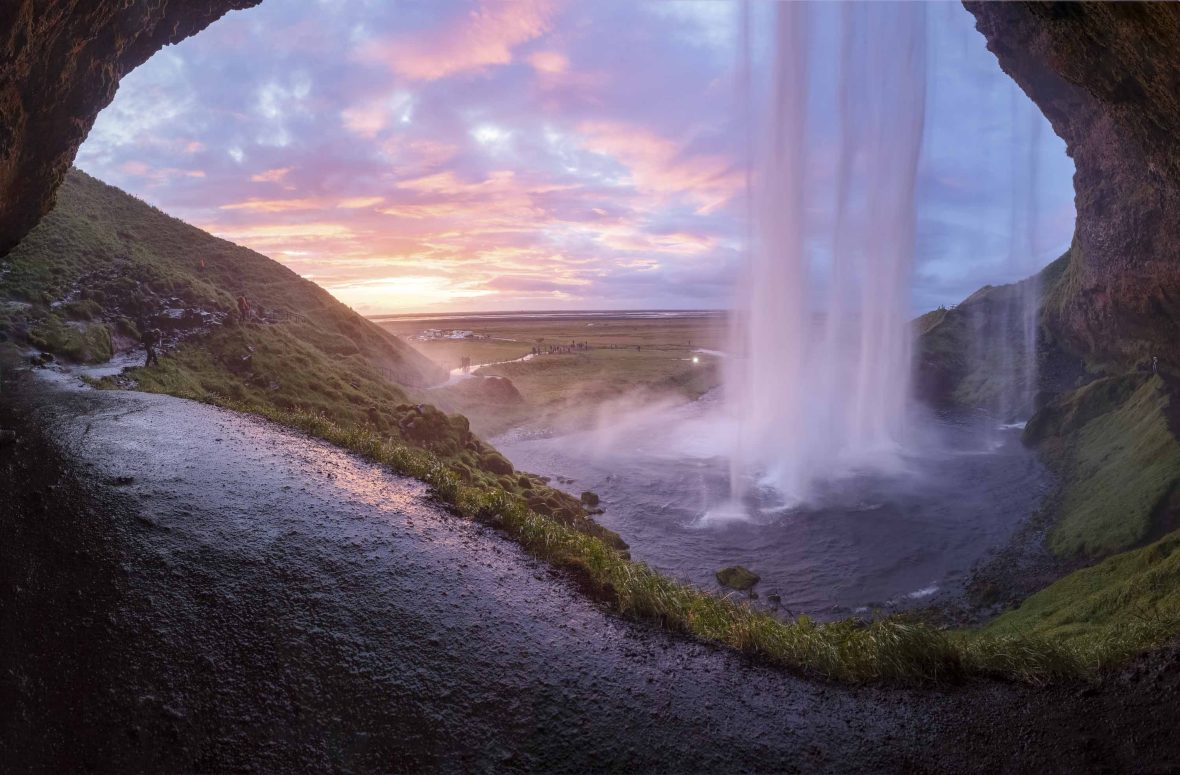 The water from a waterfall is seen cascading down through the opening of a cave. The sky is a shade of magenta.