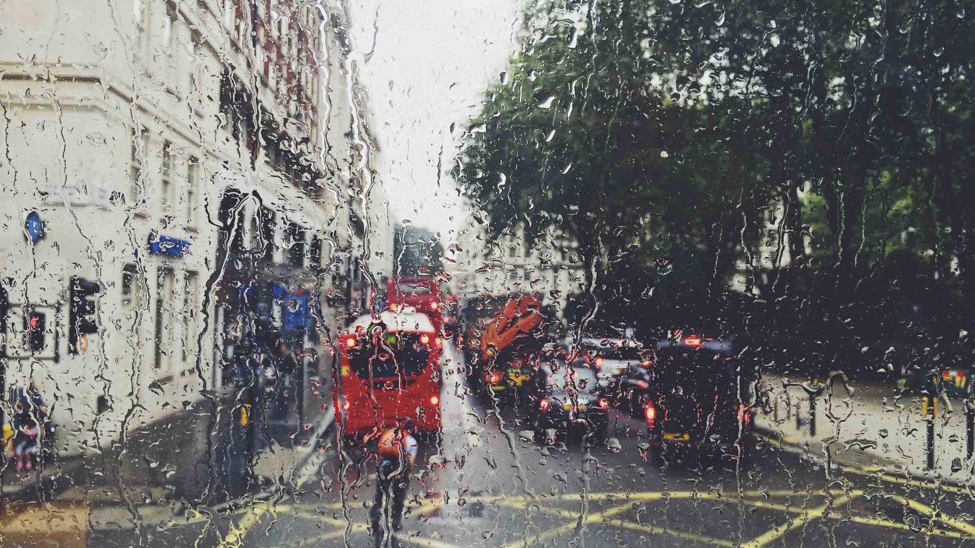 Double decker red buses are seen in the street through a wet rainy window.