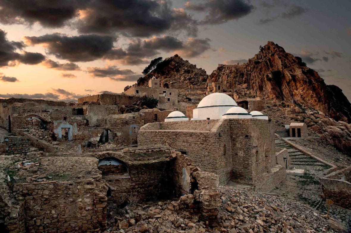 A white domed structure is visible amongst the brown ruins in an arid landscape.