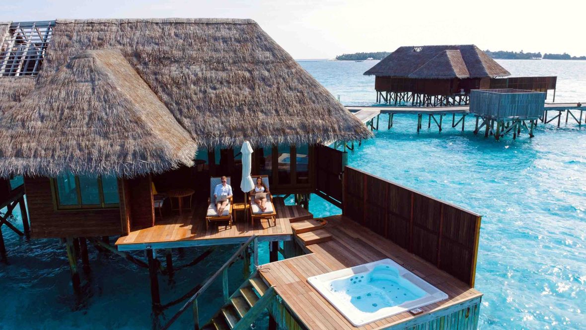 A couple work on laptops as they sit on deck chairs at luxury accomodation with a pool and clear blue water surrounding them.