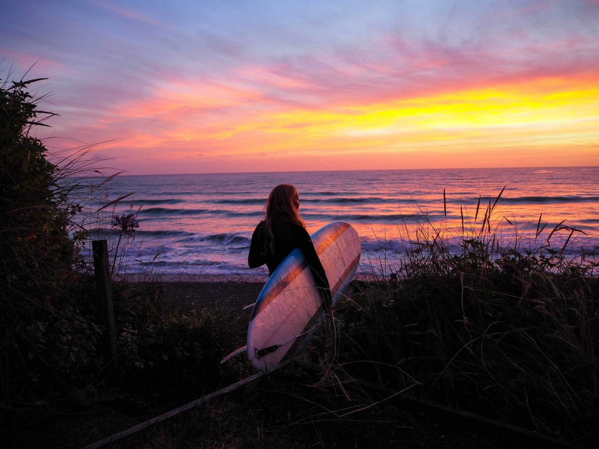 A woman holding a surfboard looks out at the water under an orange sky.