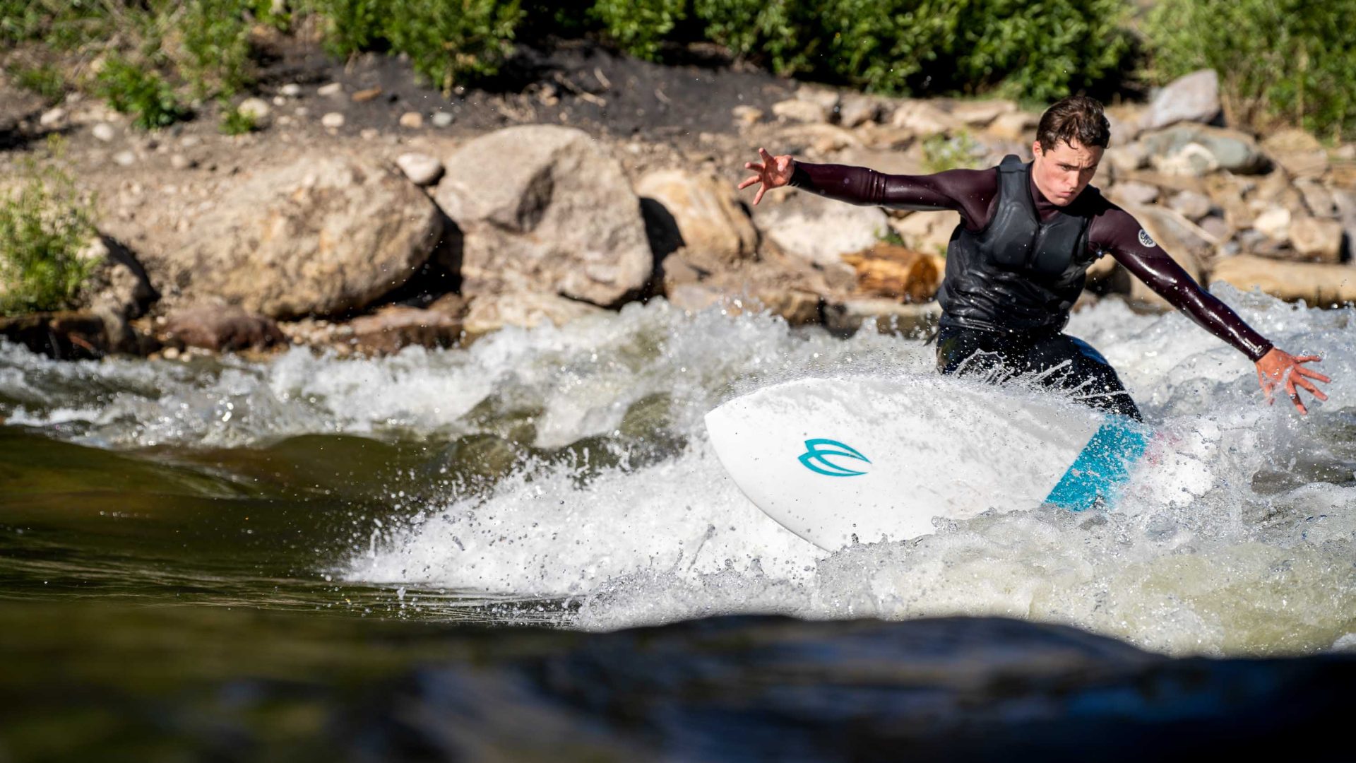A man surfs on a river. He wears a full wetsuit.