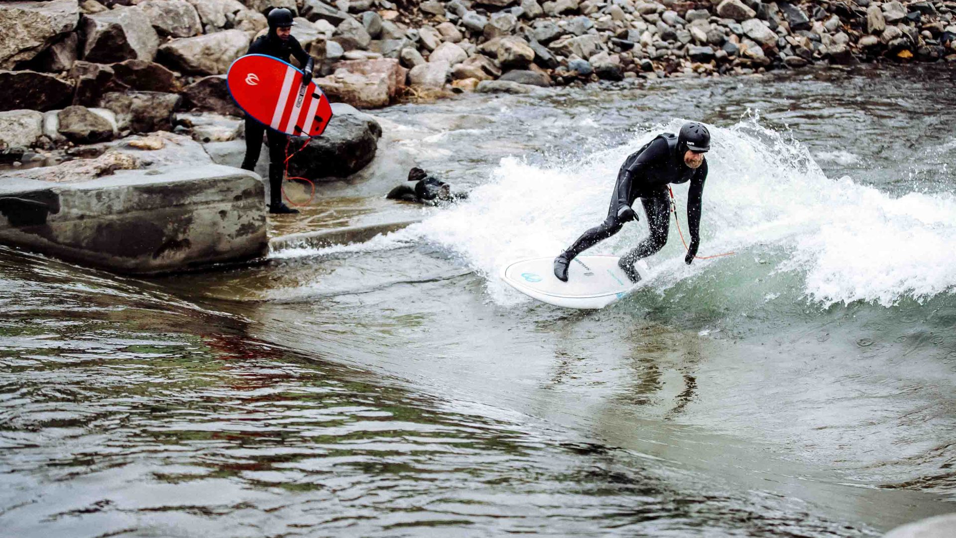 One man watches on while another surfs a river wave.