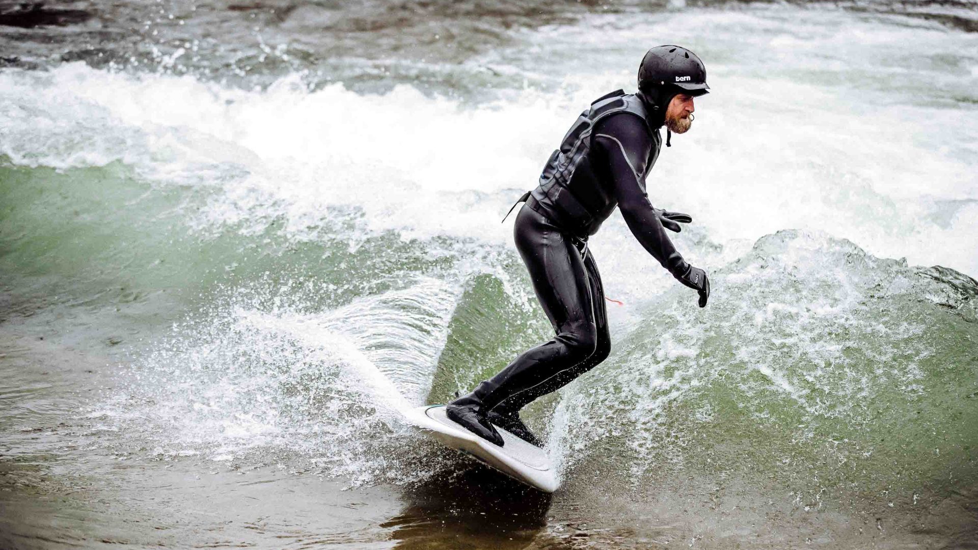 A man surfs on a river. He wears a full wetsuit and helmet.