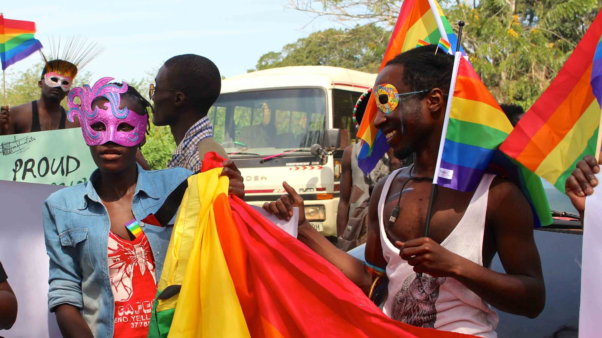 People wearing decorative masks over their eyes and holding pride flags walk in front of a white bus in a pride parade.