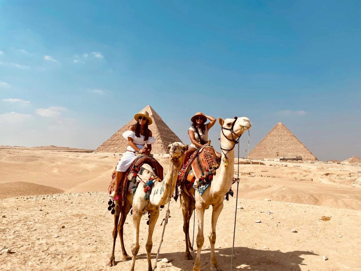 Two women ride camels in front of the pyramids in Egypt.