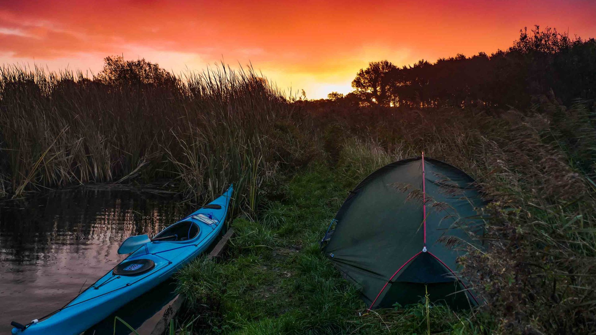 A rich orange sky, a blue kayak in the water and a green tent on the edge of the river.