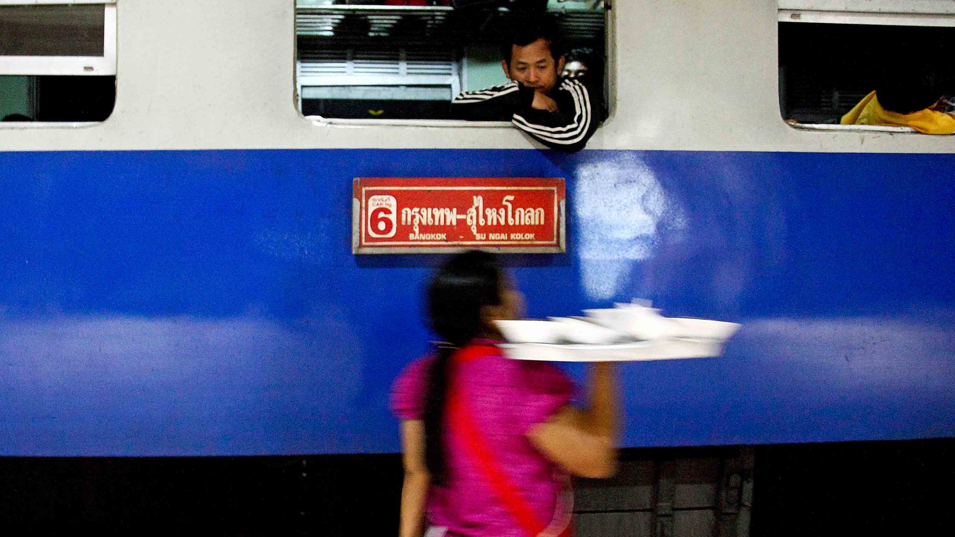 A woman selling snacks holds up a tray to a man seated in the window of a train.