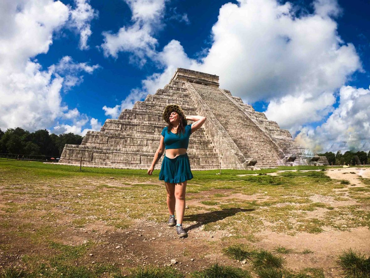 A woman in a hat stands in front of the ruins of Chichen Itza posting with her hand behind her head.
