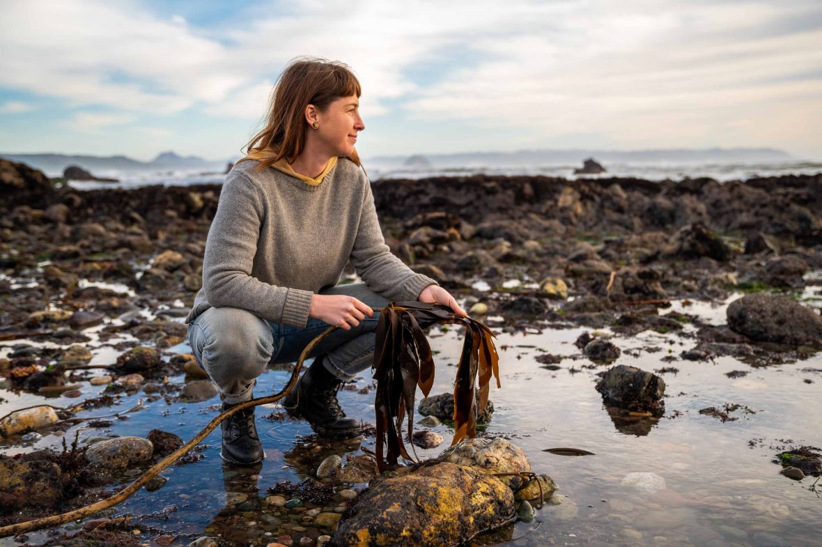 A woman looks away as she crouches on rocks holding kelp in her hand.