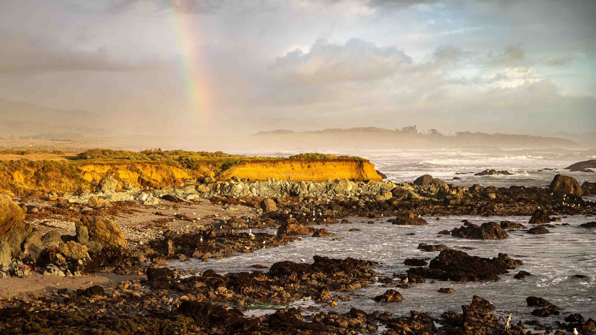 A rainbow comes down to the rocks and pools on the coast.