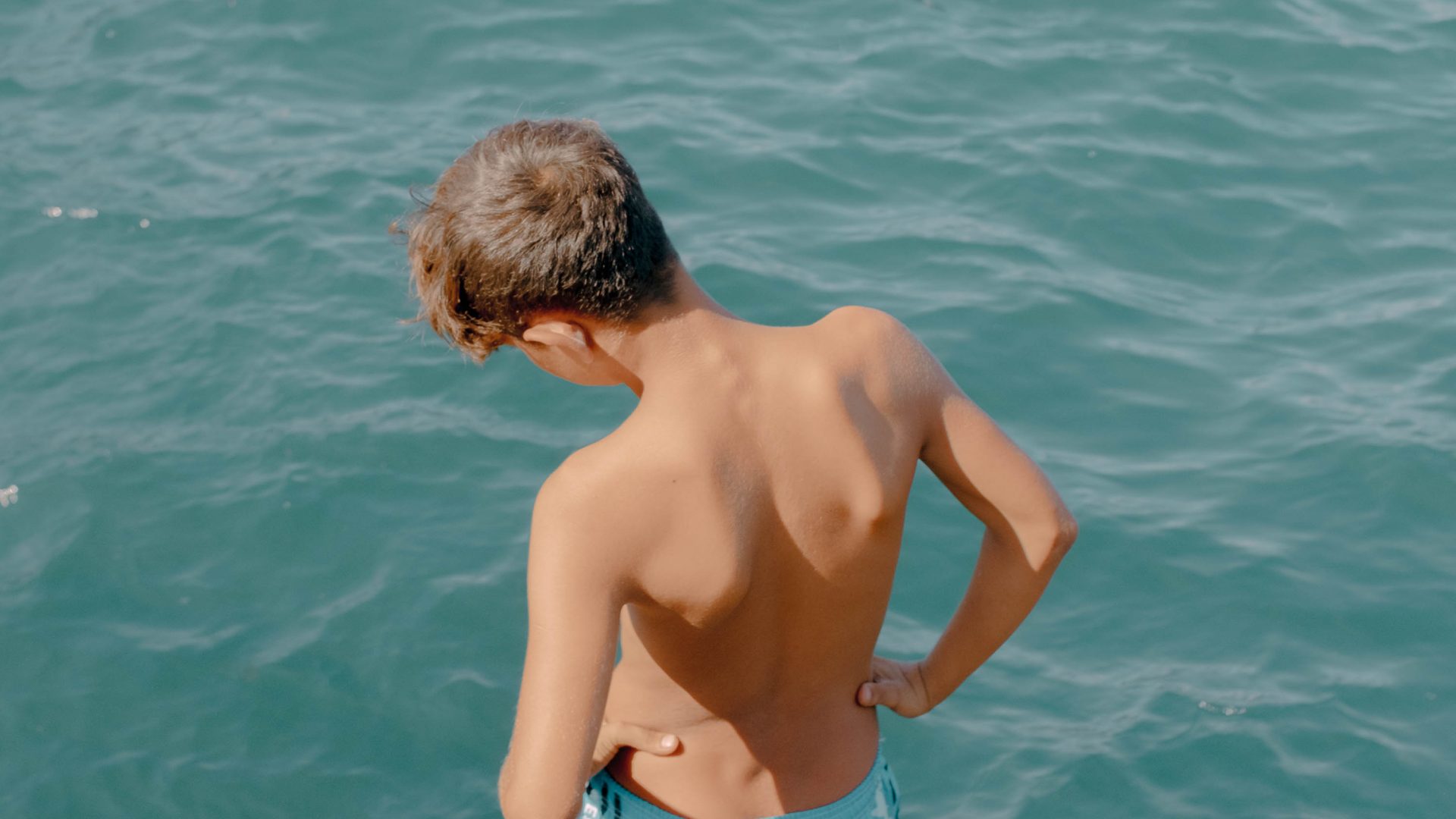A young boy in blue shorts has his back to the camera. He looks down at the blue water.