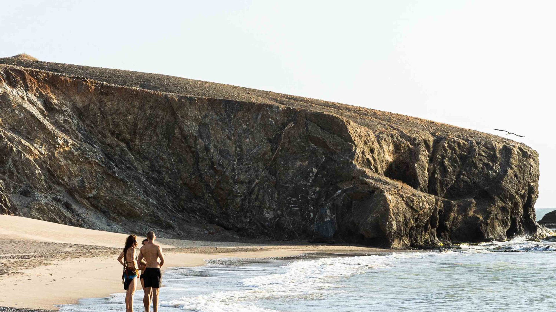 Two tourists stand by the shore at a beach fringed with cliffs.