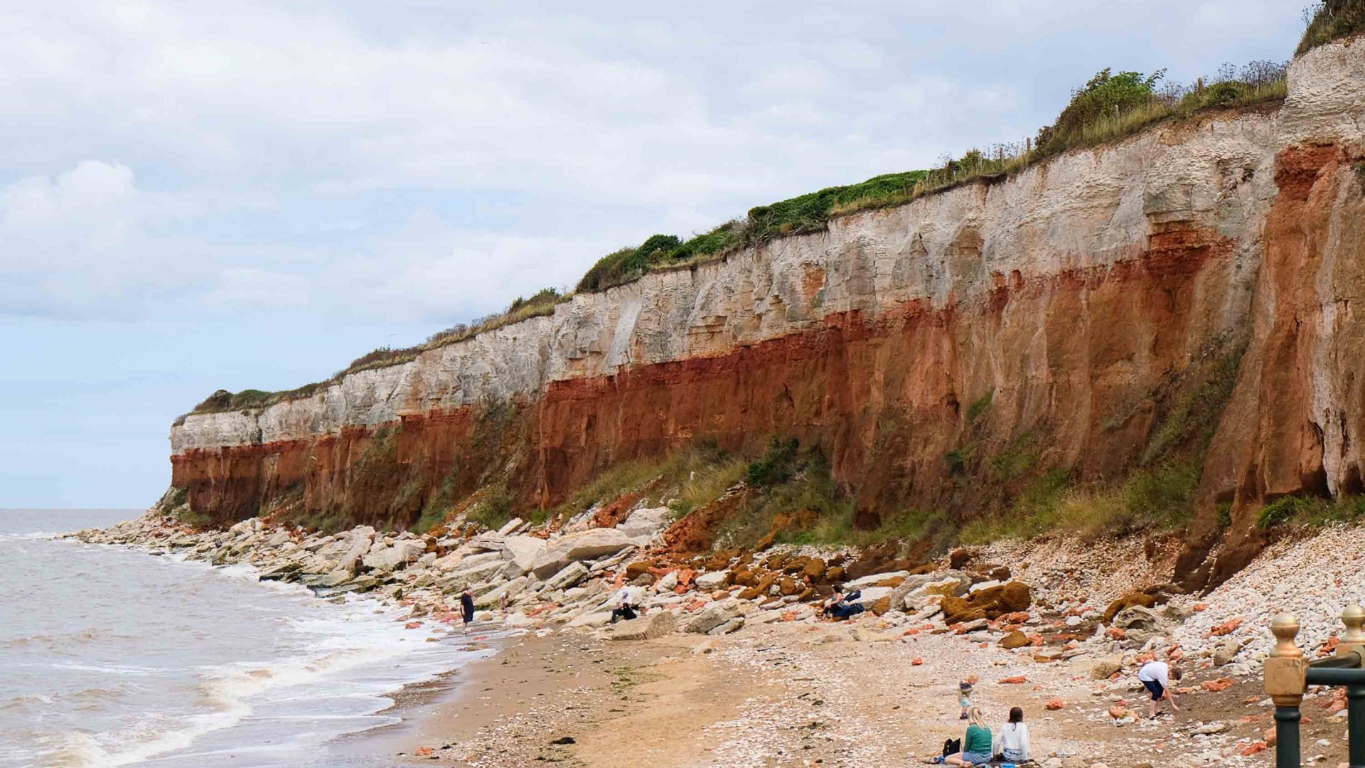 Red earth cliff face meets the ocean. A small gathering of people sit on the sand.
