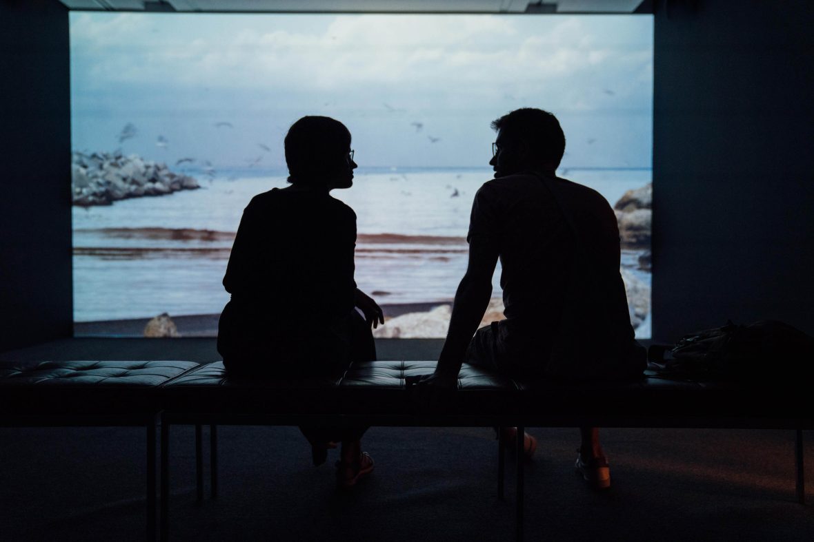 Two friends sit and talk. they are silhouetted against a window with views of water and sea birds.