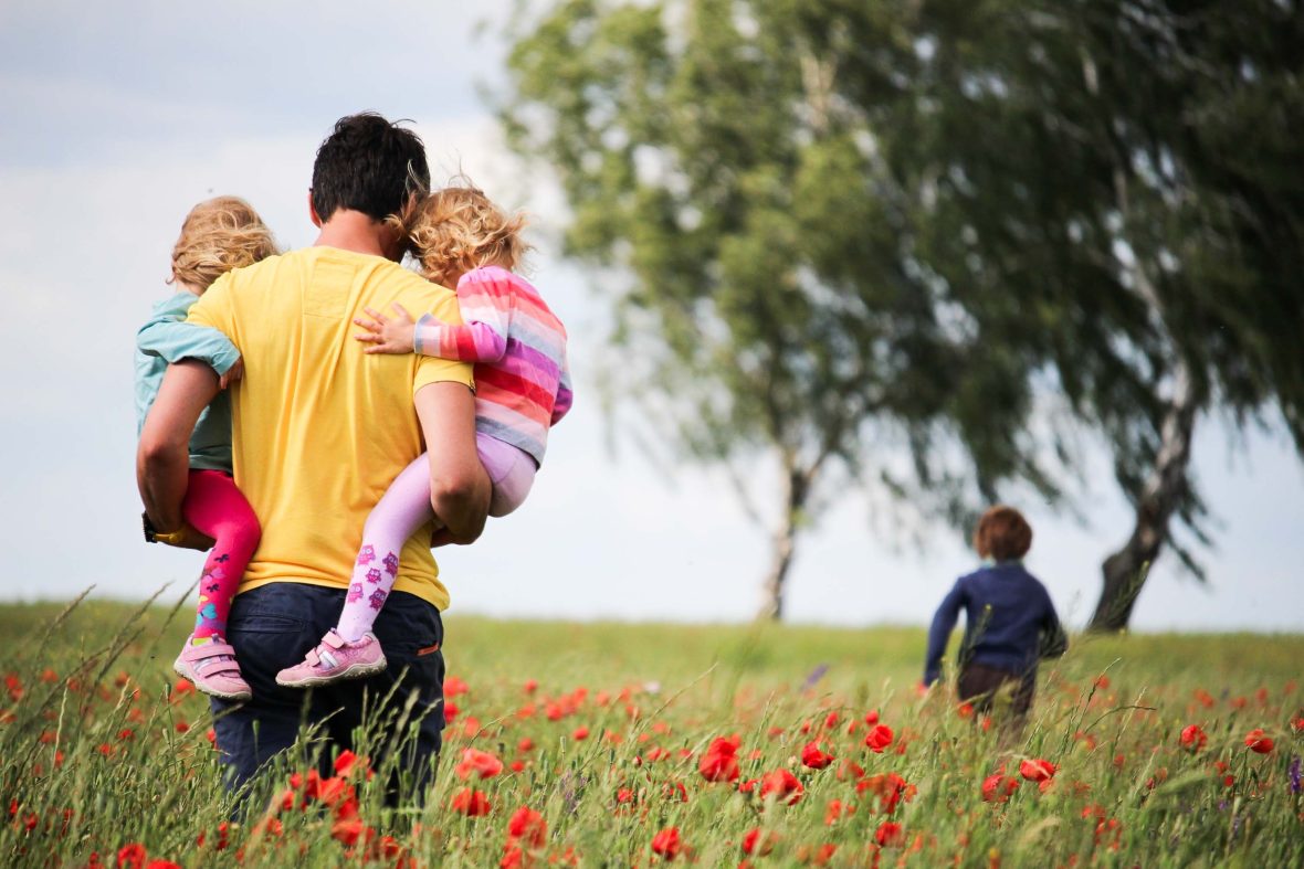 A man carries two children while a third runs ahead through a field of red flowers.