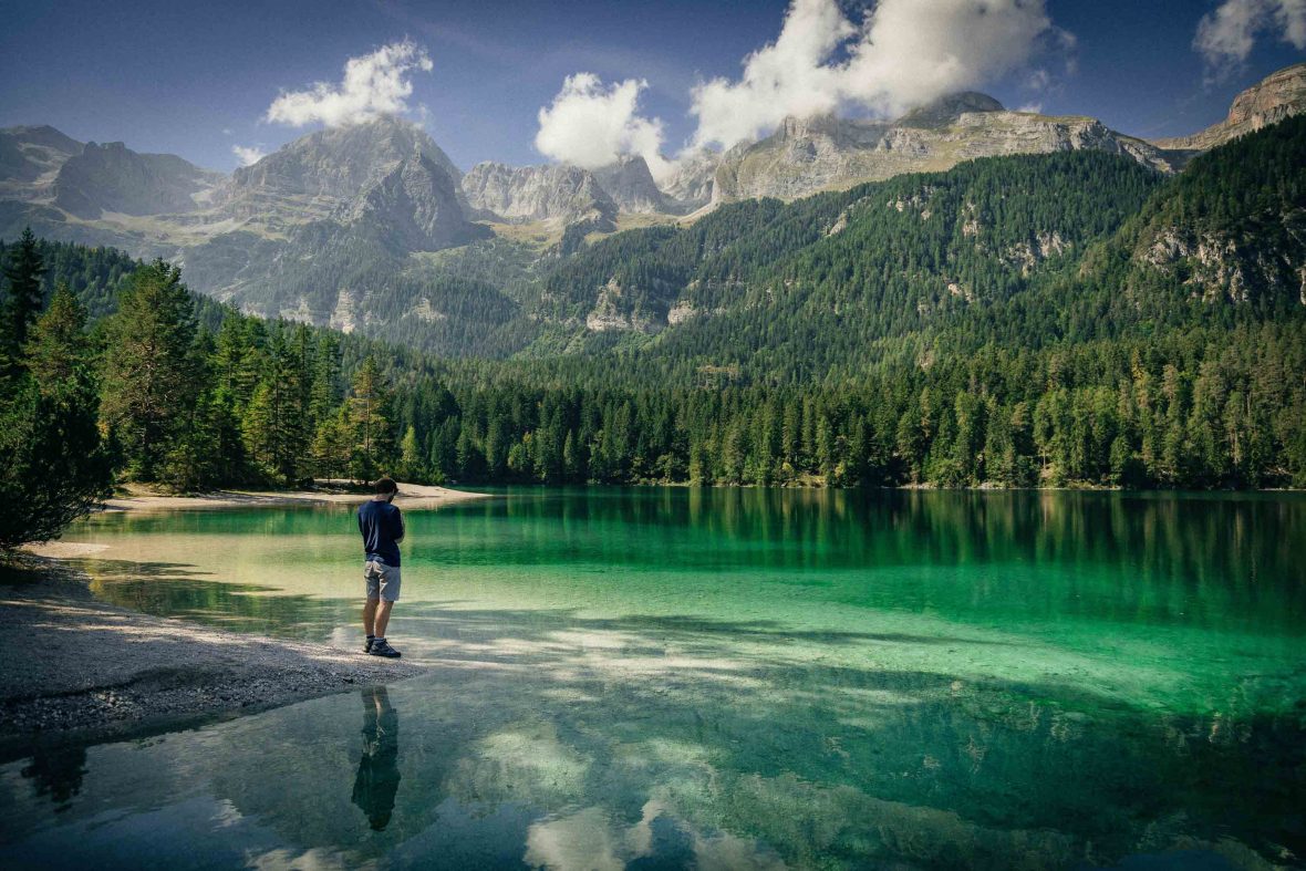 A person stands by a green lake surrounded by mountains and pine trees.