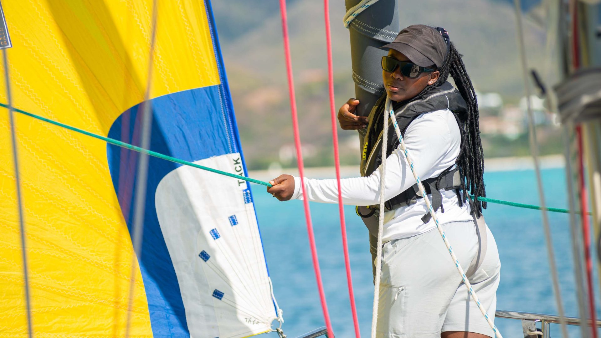 A woman stands and manages a sail which is yellow, white and blue.
