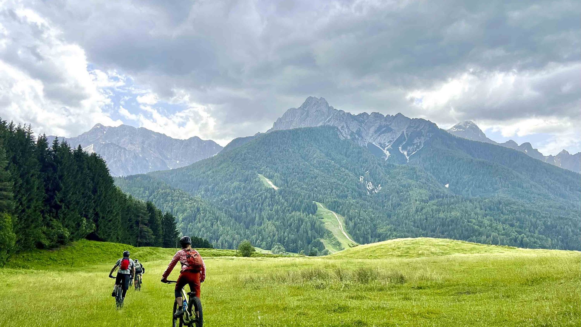 Bike riders on green pastures ride toward tall mountains.
