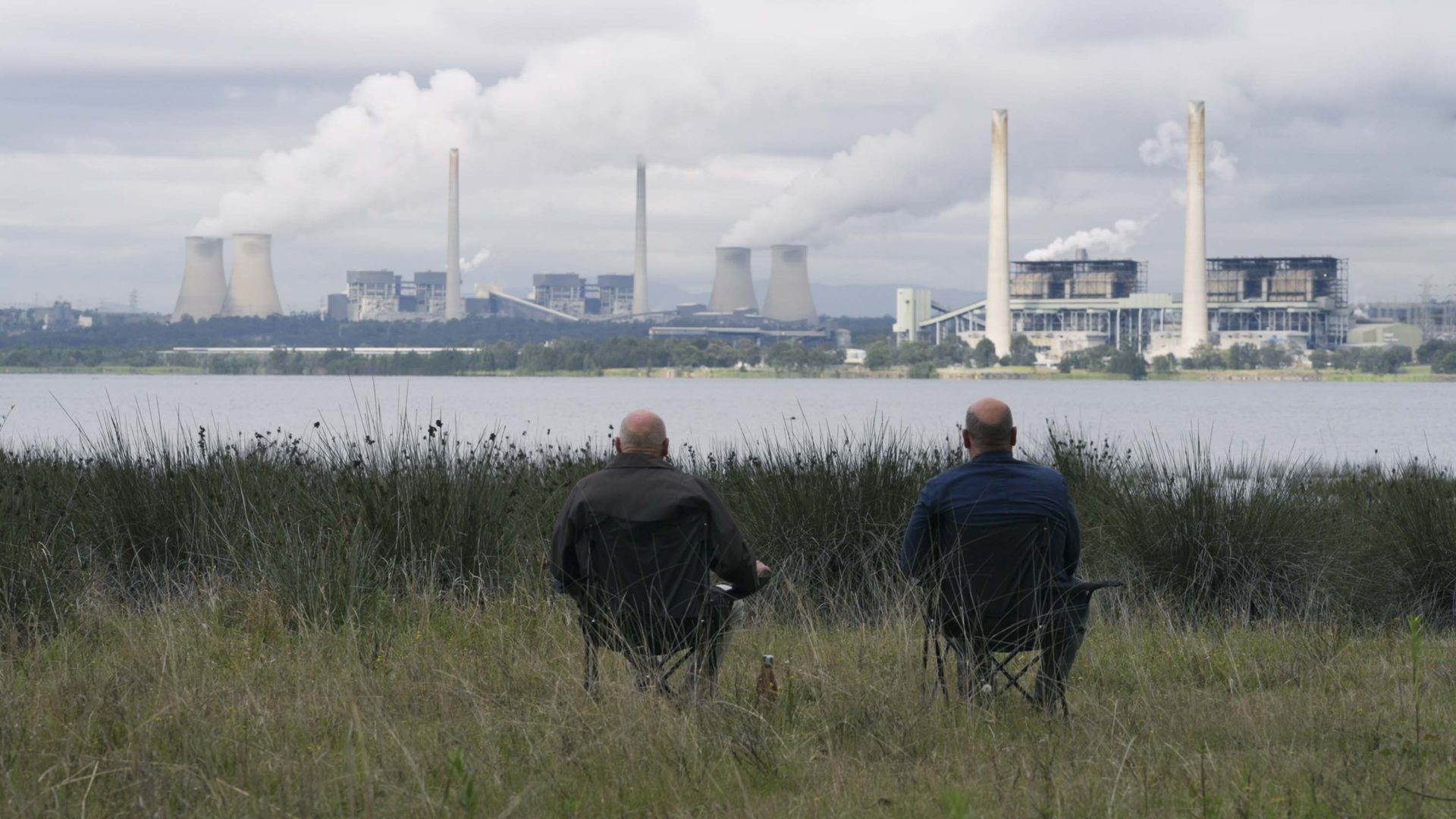 Two men sit on chairs looking over at a power station from across the water.