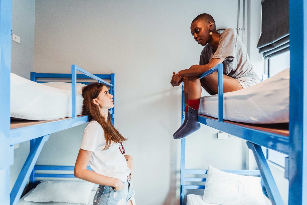 Two women sit on bunk beds at a youth hostel and talk.