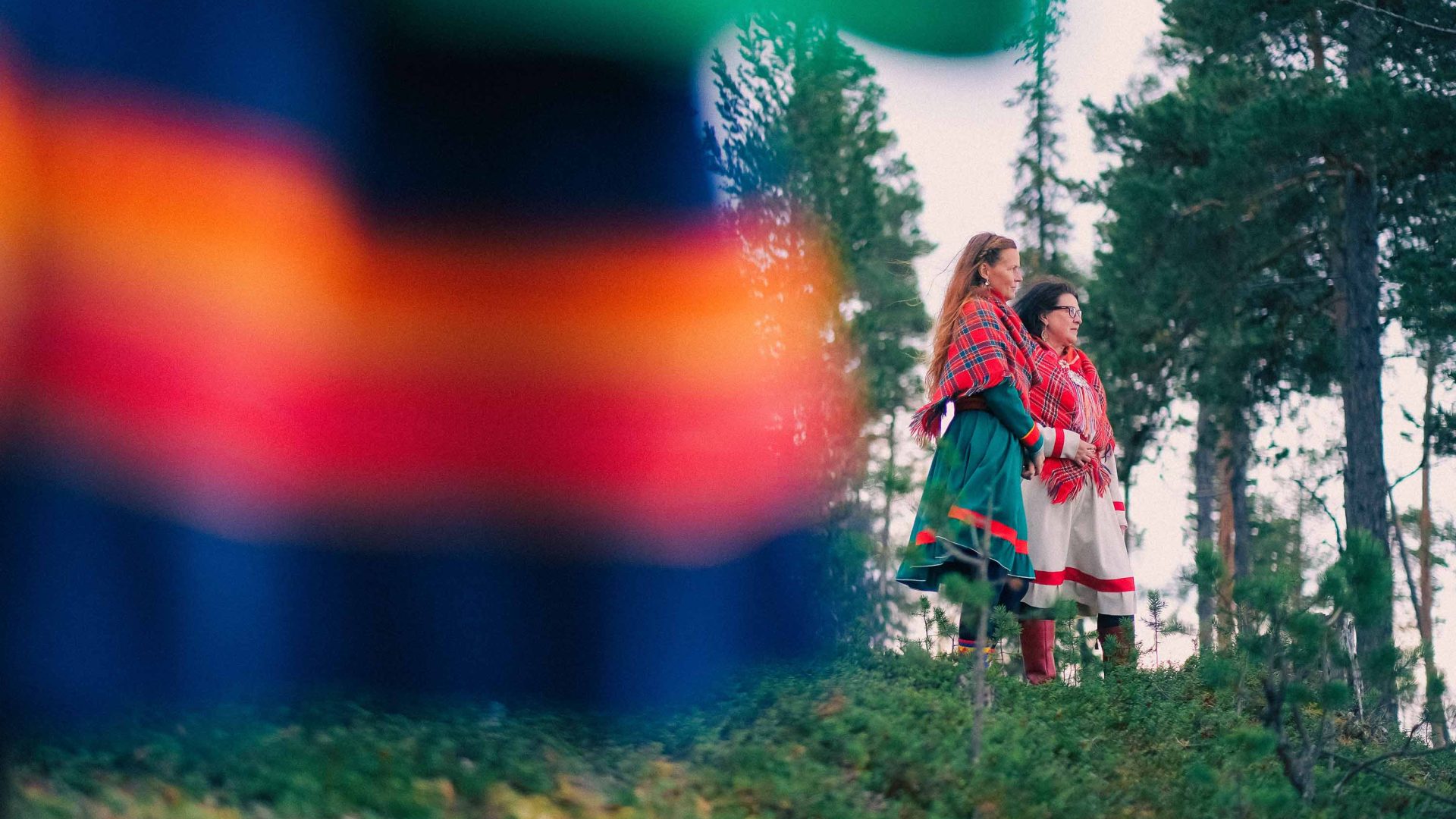 Two women look off into the distance. They are wearing colorful dresses.