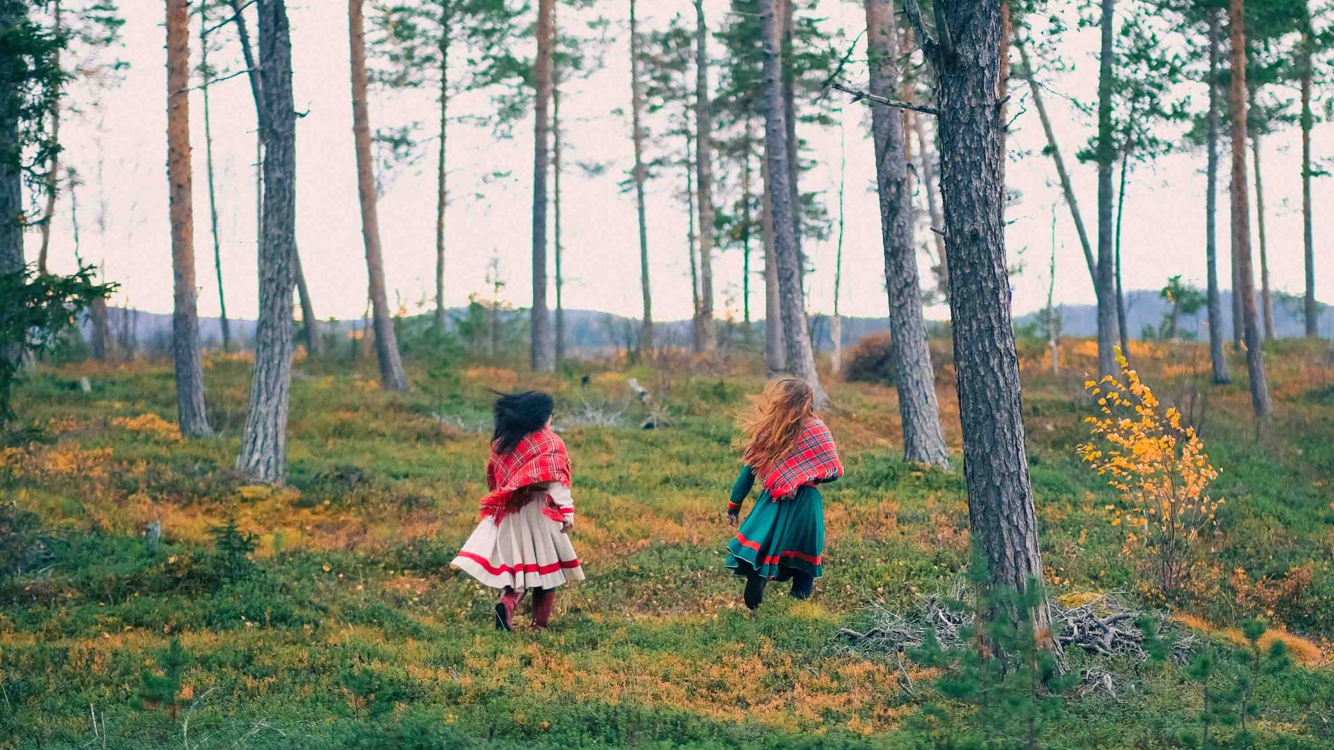 The backs of two women in colorful dresses run through forest.