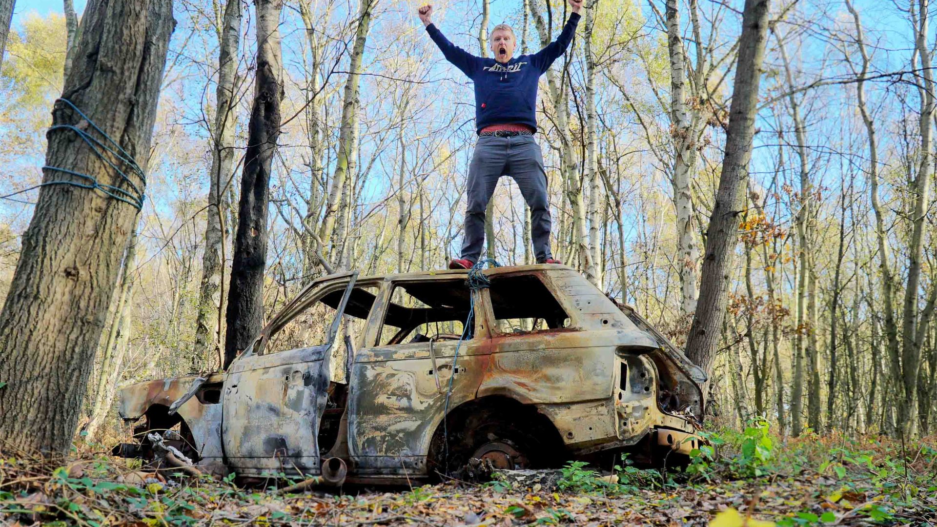 Alastair stands on top of an abandoned car in a forested area. His arms are outstretched.