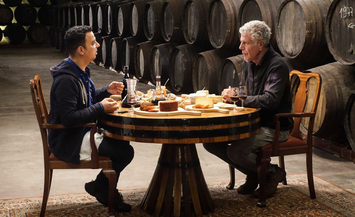 Anthony Bourdain eats with another man in Oporto, Portugal. They have a table covered in food and wine barrels behind them.