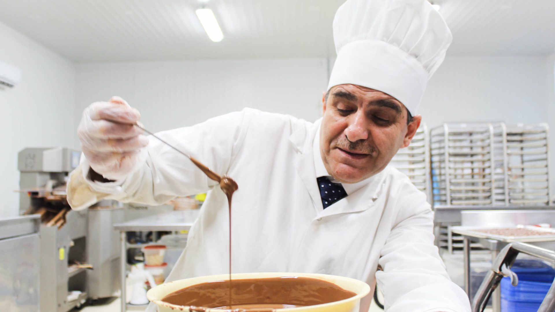 Isam Hadhad lifts up a spoon of chocolate from a big bowl. He is wearing white chef clothing.