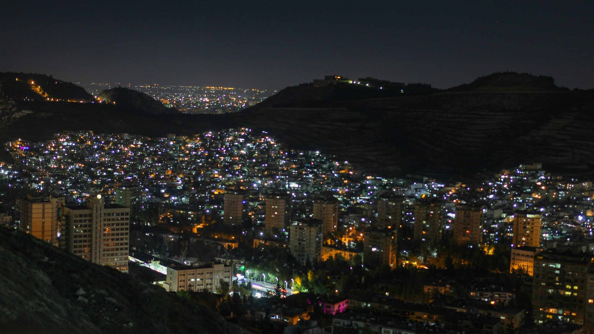 Damascus, Syria, at night. The city, lit up by lights, is surrounded by mountains.