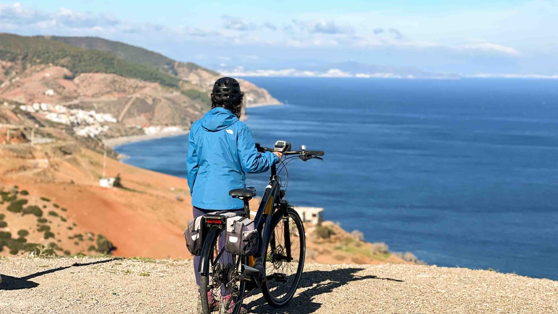 A cyclist standing on a clifftop looking at the blue sea and landscape.
