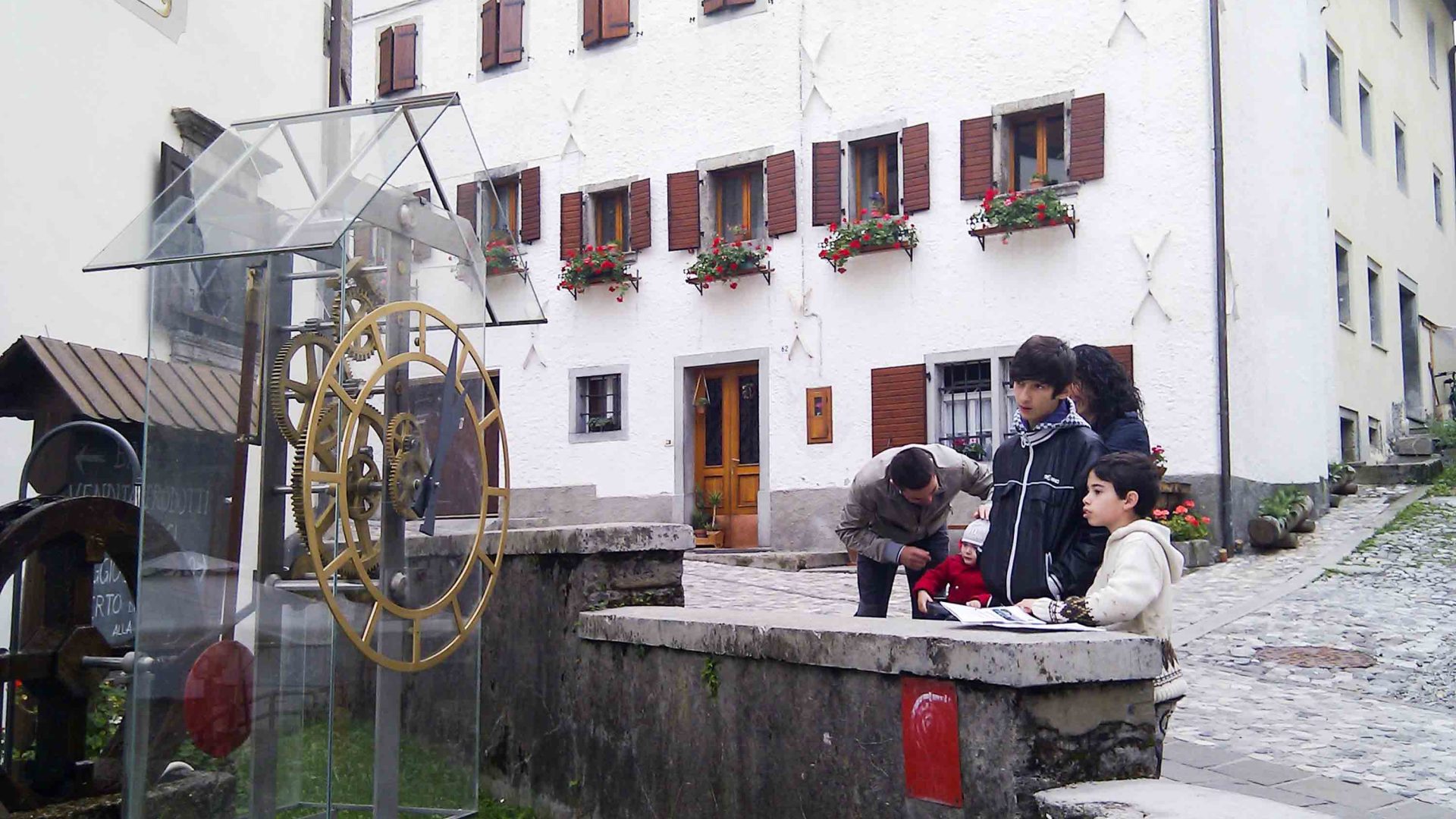 A family stops to look at a clock in Pesariis. There is a white building behind them.