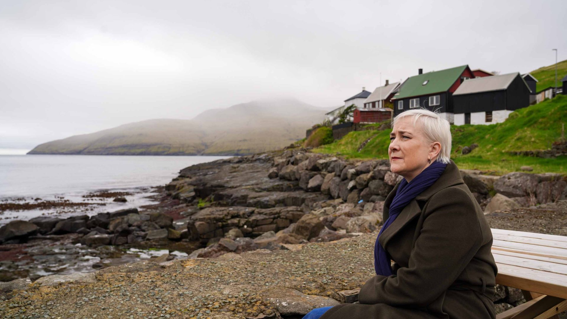 A woman with short blonde hair sits and looks out to sea.