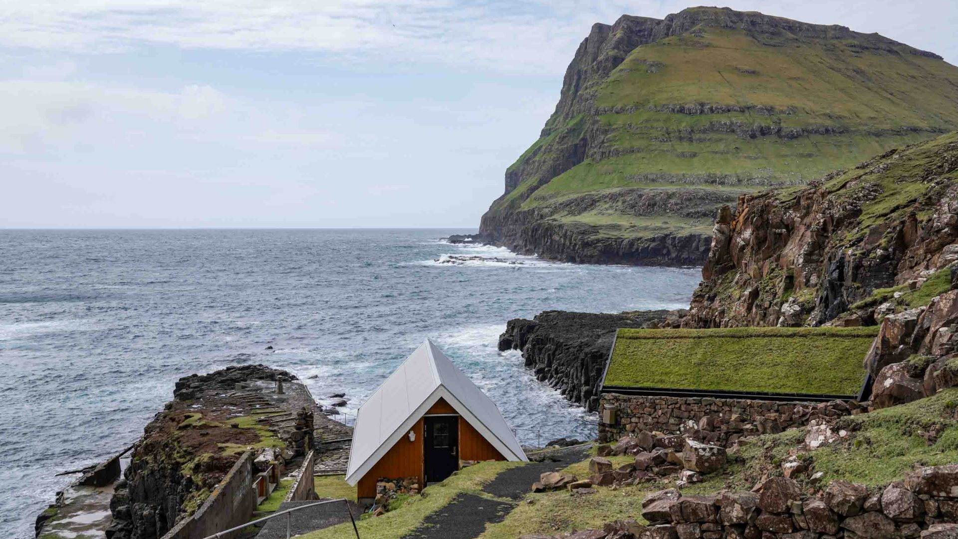 A small building with a steeped roof sits on the edge of green cliffs that drop down to the ocean.