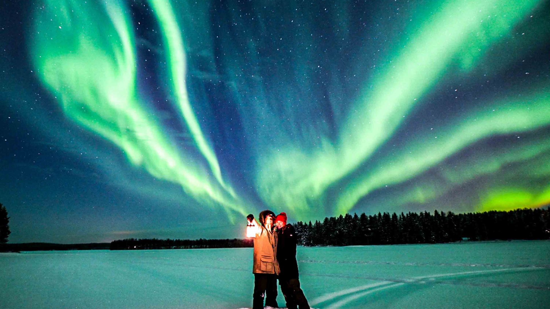A couple stand in the Arctic, under green Aurora in the sky.