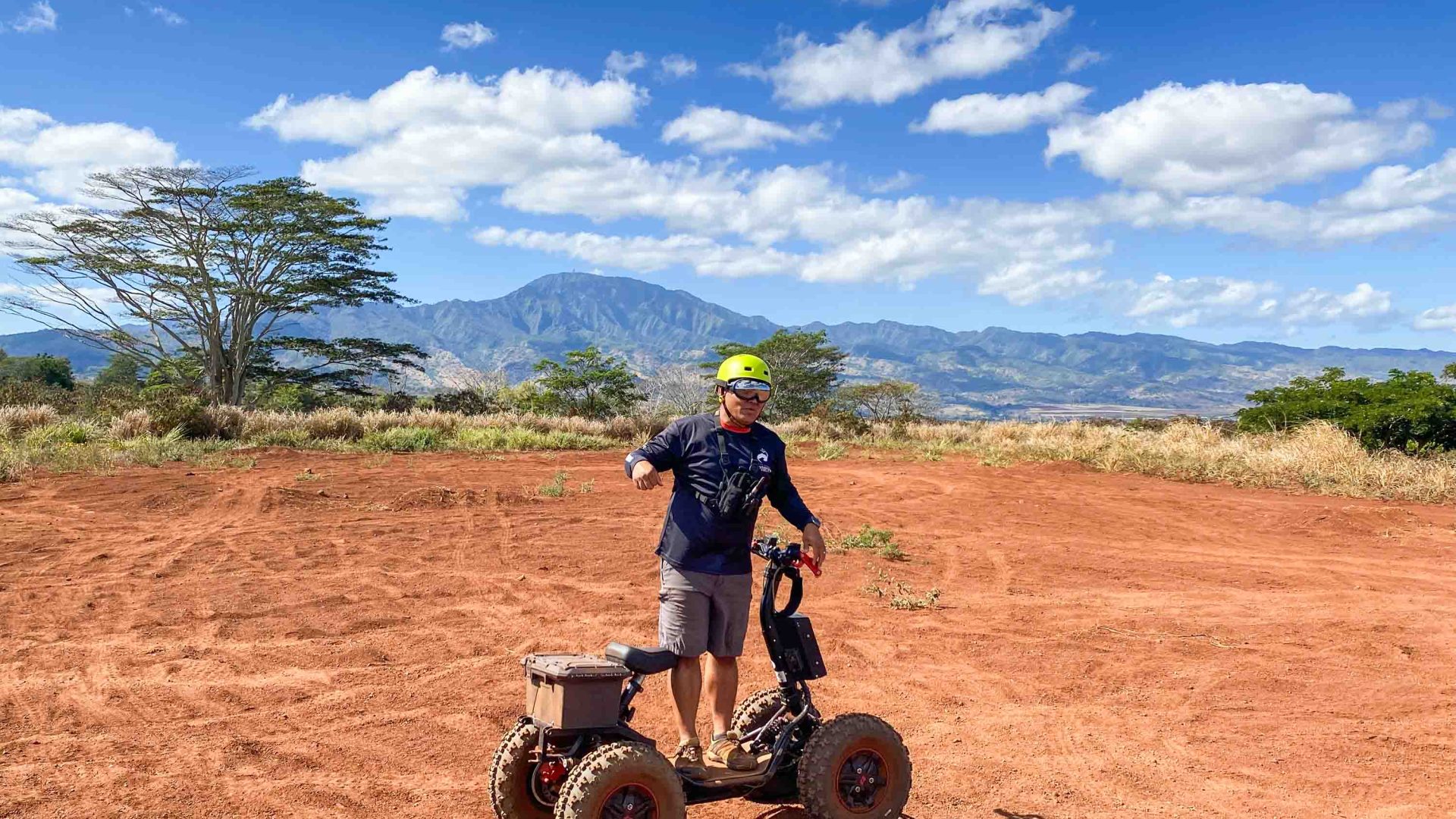 Keola stands next to his all terrain vehicle. The ground is red sand and there are mountains in the background.