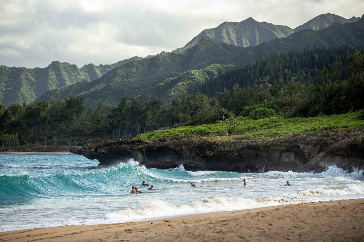 Surfers at a beach in Oahu which is backed by green mountains.