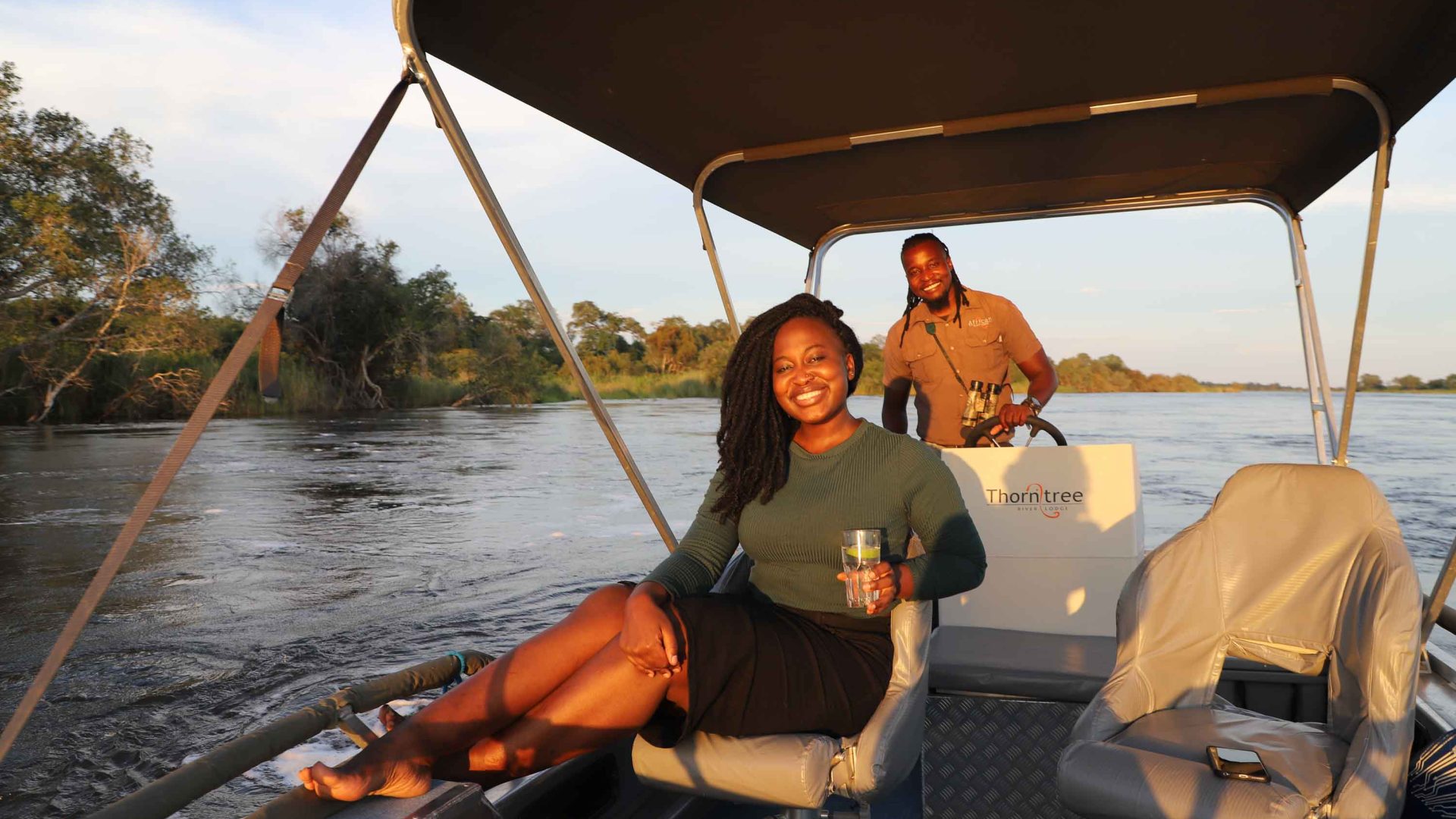 A woman enjoys a drink while on a boat in a river. The driver of the boat smiles behind her.