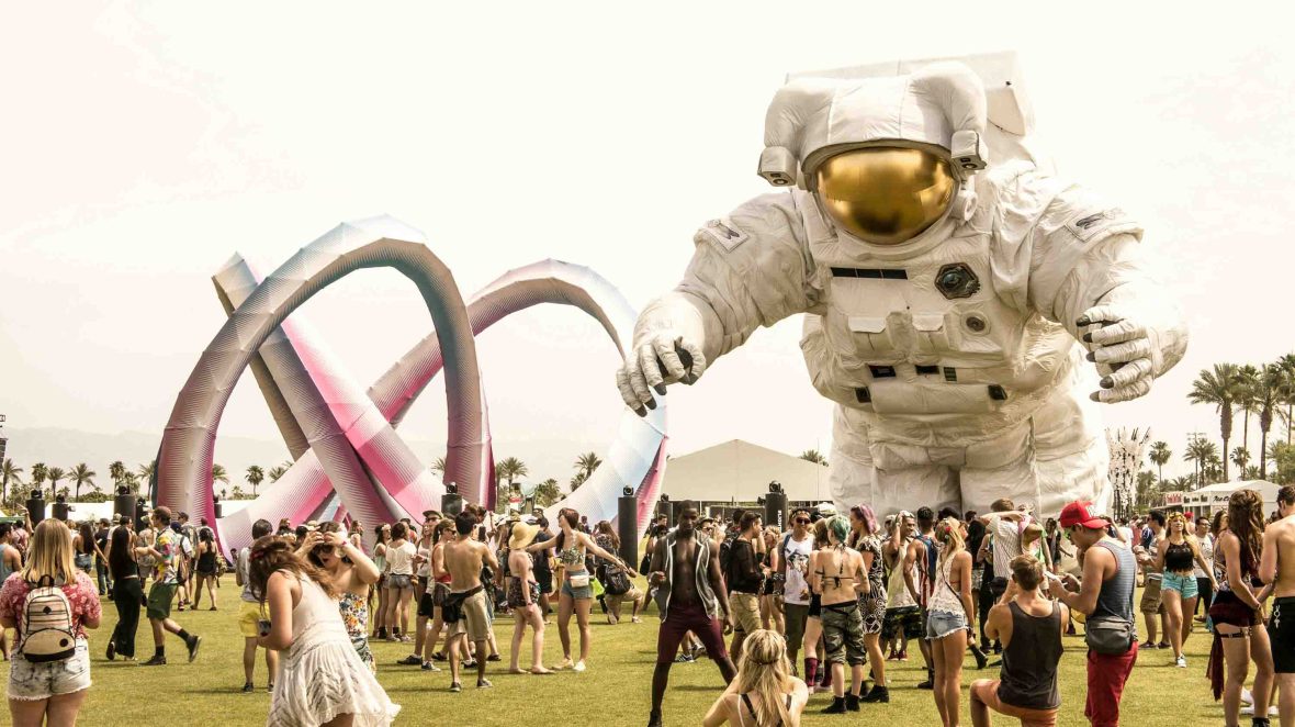 A crowd of people standing on grass, talking and dancing with a large astronaut art installation above them.