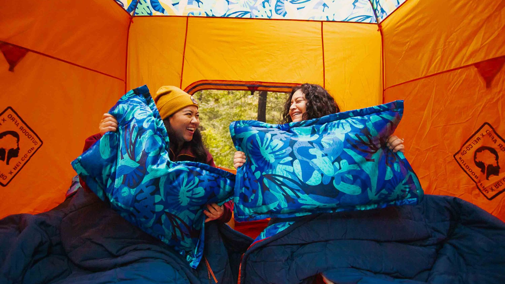 Two people in a tent hold pillows and smile at each other.