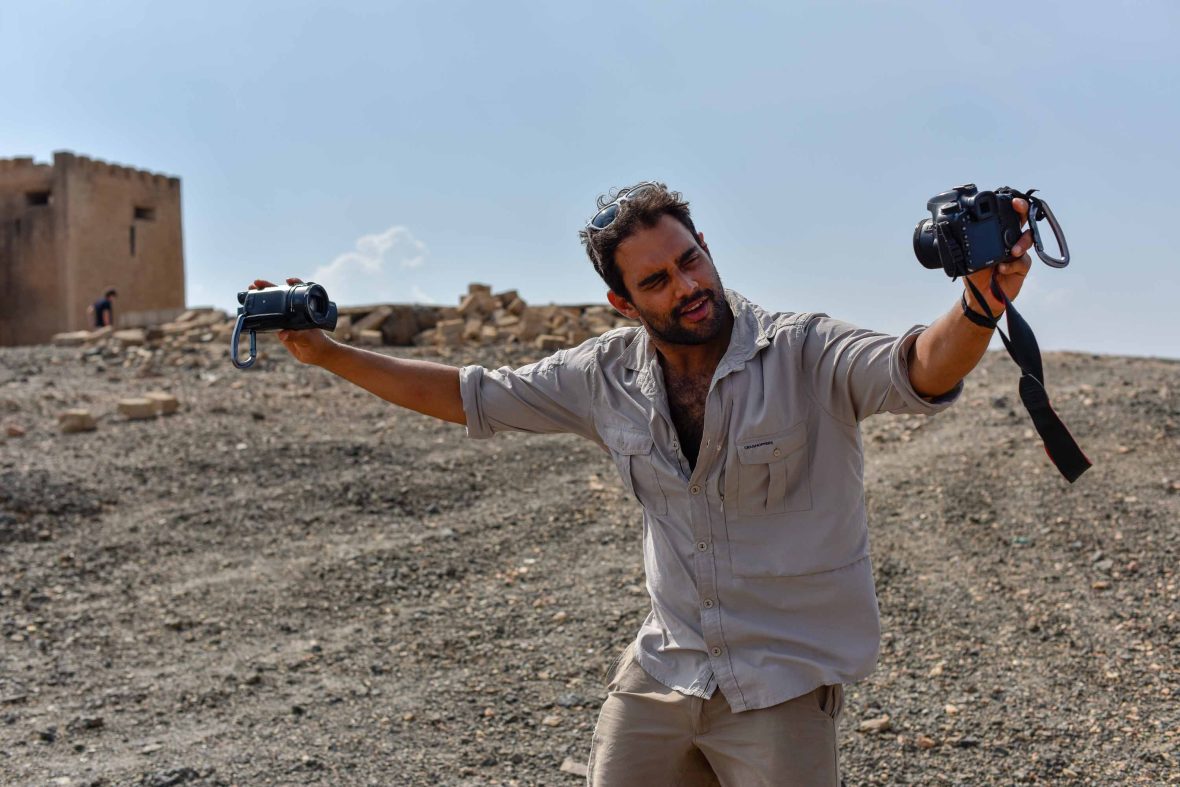 A man holds two cameras and strikes a pose in a desert environment.