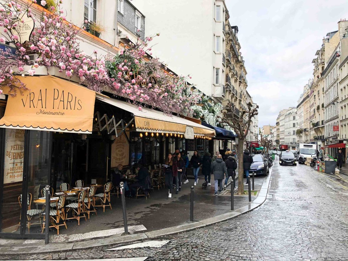 A cafe with pink flowers over the facade on the corner of a cobbled street in Paris.